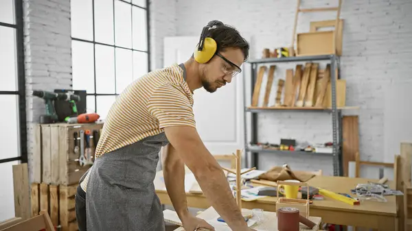 A focused hispanic man wearing ear protectors works diligently in a well-organized carpentry studio.