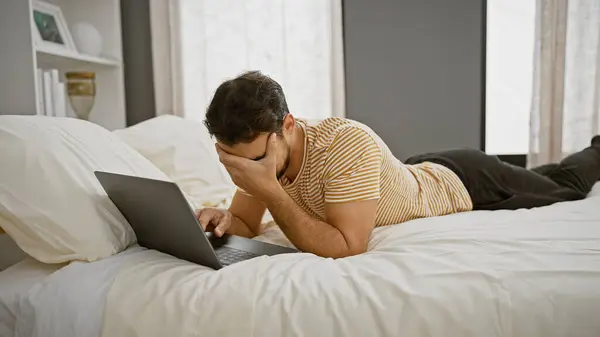 A stressed hispanic man lying on a bed using a laptop in a modern bedroom setting, exuding a sense of frustration and exhaustion.