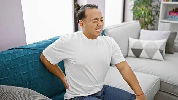 Worried young chinese man, suffering serious backache, sitting unhappily on sofa at home, showcasing the harsh indoor reality of spinal injuries.