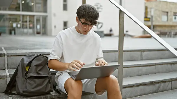 Cool hispanic teenager, a smart university student, sits on urban campus stairs, deeply engrossed in online study on his laptop. looking serious yet relaxed, living the casual city lifestyle.