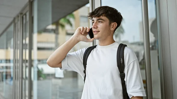 Cool young hispanic teenager studying at university, looking confident while carrying backpack and talking on his smart gadget, smiling in a casual, laid-back city-style outdoors conversation
