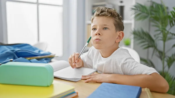 Adorable blond boy student, focused on learning, taking notes sitting at desk in indoor classroom, thinking hard on academic studies