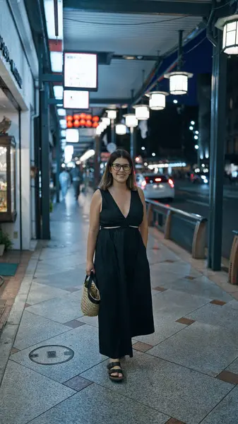 Laughing out loud, a confident beautiful hispanic woman wearing glasses stands joyfully on a street in kyoto, light-filled japanese city at night, looking positively radiant and carefree