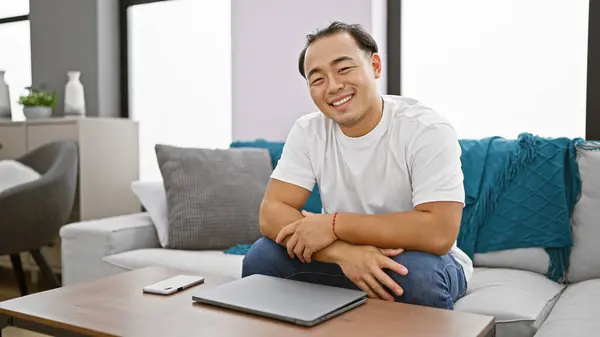 Cheerful young chinese man, devoted to his laptop, reveling in the comfort of his sofa at home in classic pigtail hairstyle