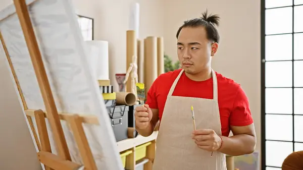 Handsome young chinese man, confident artist, drawing indoors, immersed in creativity at art studio with paintbrush in hand, adorned with traditional pigtail hairstyle & apron