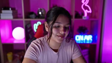 Kulaklıklı İspanyol kadın, neon ışıklarıyla aydınlatılmış canlı bir oyun odasında gülümsüyor..