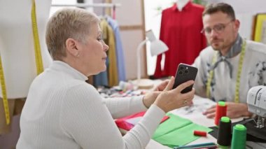 İki terzi, güneşli bir atölyede renkli kumaşlarla işbirliği içinde çalışıyor. Biri akıllı telefon kullanıyor..