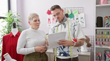 İki terzi, bir erkek ve bir kadın, eskizler ve kumaşlarla donanımlı bir terzide işbirliği yapıyorlar.