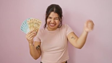 Heyecanlı genç İspanyol kadın zaferi kutluyor! Brezilya banknotlarını pembe izole edilmiş arka planda tutuyor. Güzel ve gururlu, başarısıyla, zaferiyle neşe ve güven dolu.