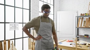 Gözlüklü ve önlüklü, kendinden emin Afro-Amerikalı bir adam, elinde matkapla bir marangoz atölyesinde poz veriyor..