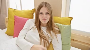 Feisty young woman in pyjama flips the bird in bedroom, showing off a rude 'fuck you' expression sitting comfortably on her bed