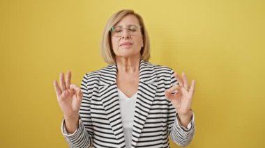 Güzel, orta yaşlı sarışın yoga pozunda meditasyon yapıyor, gözleri kapalı, çizgili ceket giyiyor. İzole edilmiş sarı arkaplanda sağIık kavramı.