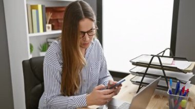 Modern bir ofis masasında akıllı telefon ve belgelerle çalışan odaklanmış gözlüklü bir kadın.