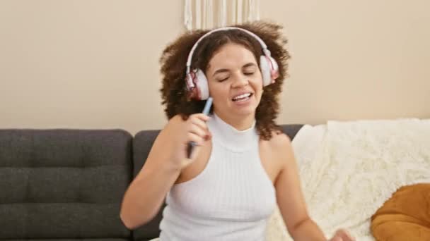 一位卷发的年轻惊慌失措的女人喜欢用耳机听音乐 在家用手机唱歌 — 图库视频影像