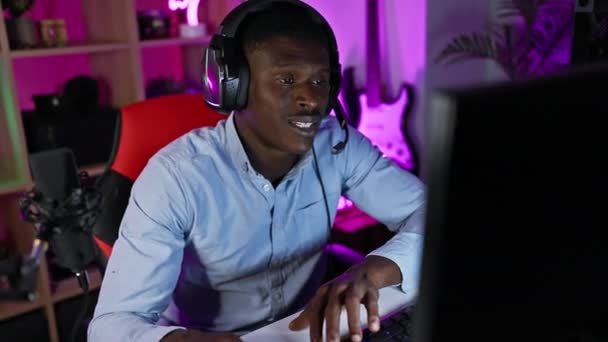 在霓虹灯照明的游戏室里 一个戴着耳机的笑着的非洲男人正在进行在线对话 — 图库视频影像