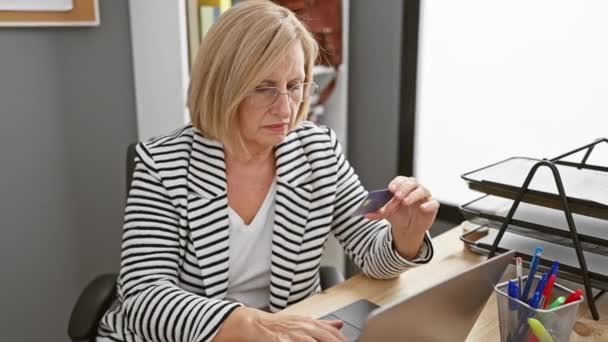 Mature Woman Examining Credit Card Skeptically Office Desk Laptop Pens — Stock Video