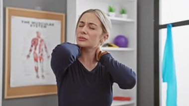Sarışın bir kadın rehabilitasyon kliniğinde arka planda kas sistemi posteriyle boyun ağrısı çekiyor.