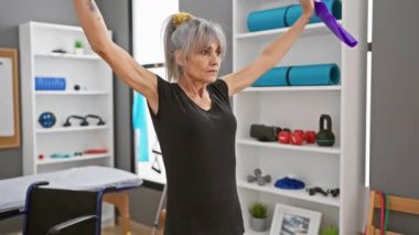 Olgun kadın, iyi donanımlı bir terapi odasında direniş bantlarıyla egzersiz yapıyor, sağlık ve rehabilitasyon gösteriyor..