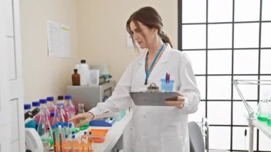 Laboratuvarda yetişkin bir kadın bilim adamı elinde bir pano tutarken test tüplerini inceliyor, klinik ve araştırma atmosferi sağlıyor..