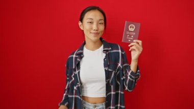 Neşeli genç Çinli kadın, Çin pasaportuyla geziyor. Heyecan verici bir şekilde, kızıl izole edilmiş bir arka planda, neşe ve kendine güvenle işaret ediyor.!