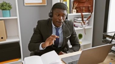 Kulaklık takan odaklanmış Afrikalı bir adam modern ofis ortamında video görüşmesi için dizüstü bilgisayar kullanıyor..