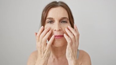 Orta yaşlı bir kadın beyaz bir arkaplanda yüzünü inceliyor. Güzellik ve cilt bakımını resmediyor..