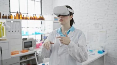 Laboratuvar önlüklü genç bir bilim kadını modern bir laboratuvar ortamında sanal gerçeklik gözlüğü kullanıyor.