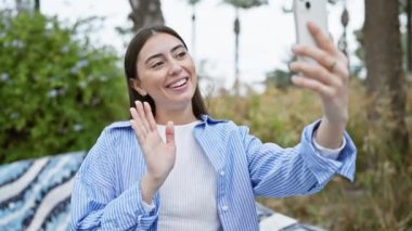 Çizgili tişörtlü, gülümseyen genç İspanyol kadın açık hava parkında akıllı telefonuyla görüntülü görüşme yapıyor.