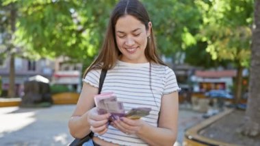 Güneşli bir parkta Endonezya parası sayan mutlu genç bir kadın, boş zamanlarını ve seyahatlerini resmediyor..