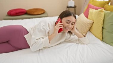 Genç İspanyol kadın renkli bir yatak odasında akıllı bir telefonla konuşurken gülümsüyor, rahat ve rahat bir anı yakalıyor..