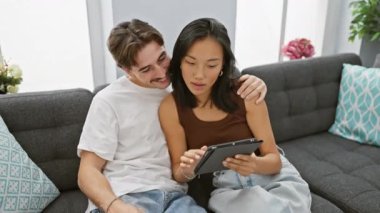 Modern oturma odasındaki rahat koltukta tablet kullanarak gülümseyen ırklar arası bir çift.