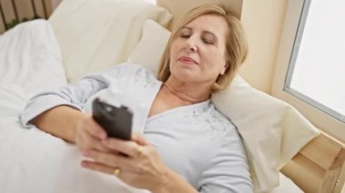 Olgun bir kadın yatak odasında akıllı telefonuyla rahatlıyor. Rahatlık, rahatlık ve modern bir bağ sergiliyor..