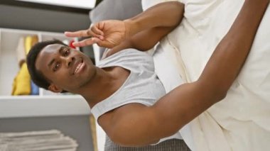 Neşeli, Afro-Amerikalı, spor pijamalı, iki parmaklı zafer işareti gösteren genç bir adam. Başarının gülümseyen bir örneği, rahat yatak odasında uzanıyor..