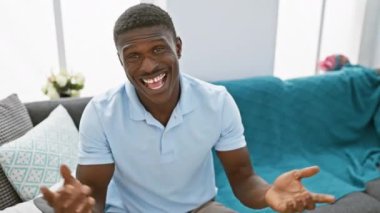 Mavi gömlekli gülümseyen Afrikalı adam modern evdeki gri koltukta oturuyor.