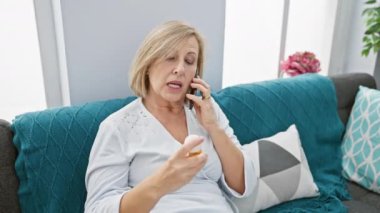 Endişeli, olgun, sarışın bir kadın elinde ilaç şişesiyle cep telefonuyla konuşuyor.