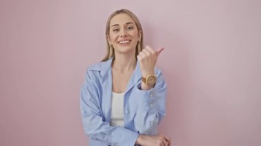 Gülümseyen genç sarışın kadın, pembe izole bir arka plan üzerinde heyecanlı bir şekilde yan tarafı işaret ediyor. Tişört giyip sevinçle ışıldıyor, pozitif titreşimleri simgeliyor.