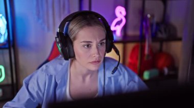 Geceleri canlı bir oyun odasında kulaklık takan, odaklanmış sarışın bir kadın..