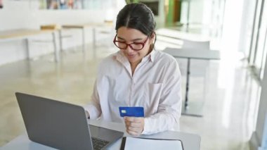 Modern ofis ortamında dizüstü bilgisayar üzerinde çalışırken elinde kredi kartı tutan İspanyol kadın.