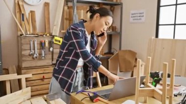 Asyalı kadın, bilgisayar ve telefon kullanırken ahşap atölyesinde birden fazla iş yapıyor..