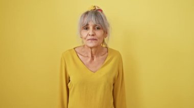 Orta yaşlı neşeli bir kadın, kameraya seksi bir şekilde göz kırpıyor. Mutlu yüz, atkuyruklu gri saçlar, izole edilmiş sarı arka planda..