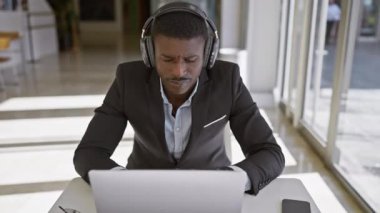 Modern bir ofiste dizüstü bilgisayar kullanan, kulaklık takan, odaklanmış Afro-Amerikan bir adam..