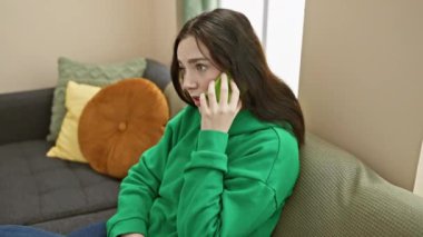 Yeşil kazaklı beyaz bir kadın oturma odasından neşeli bir telefon görüşmesi yapıyor..