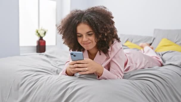 パジャマのカーリーヘアの陽気なヒスパニック女性は ベッドルームベッドに横たわりながらスマートフォンに幸せにテキストを送り 親指を横に振りかけ 開いた口の喜びの笑顔をスポーツ — ストック動画