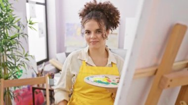 Stüdyoda oturan neşeli, kıvırcık saçlı İspanyol kadın sanatçı, parmaklarıyla işaret veriyor, parmak deliğinden bakıyor, neşeyle gülümsüyor.