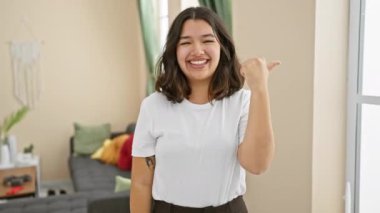 Şen şakrak, genç İspanyol bir kadın rahat oturma odasında duruyor, ağzı açık bir gülümseme takınıyor, başparmağını kenarı işaret ediyor. Saf neşe, pozitiflik ve güven yayar..