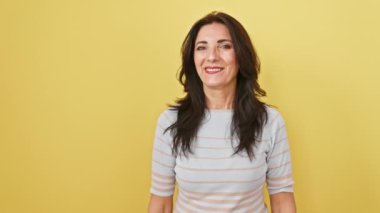 Neşeli, olgun İspanyol kadın iki elini yana doğru uzatıyor, gülümsüyor ve sarı izole bir arka plana bakıyor..