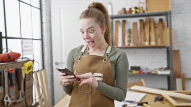 Bir atölyede akıllı telefon kullanan neşeli bir kadın, ahşap işçiliği aletleriyle çevrili.