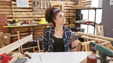 Kıvırcık saçlı genç bir kadın, ahşap ve aletlerle çevrili bir marangoz atölyesinde tableti inceliyor..