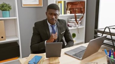 Modern ofis içinde dizüstü bilgisayar ve tabletle çalışan takım elbiseli yakışıklı bir zenci.