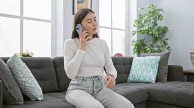 Modern bir oturma odasında akıllı telefondan konuşan İspanyol kadın zamanı kontrol ediyor..